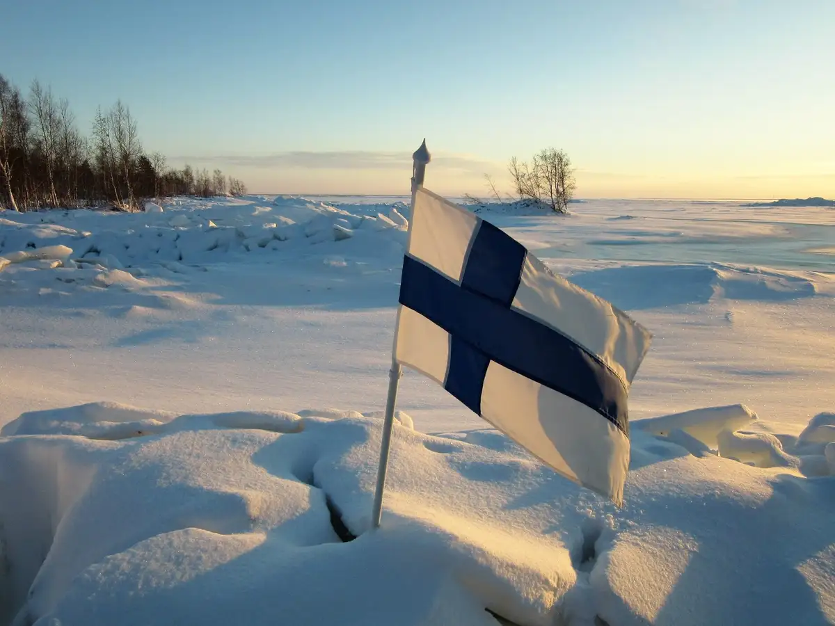 A Finnish flag stands in deep snow on a frozen, icy landscape under a clear sky, with text overlaid that reads 'finland is…