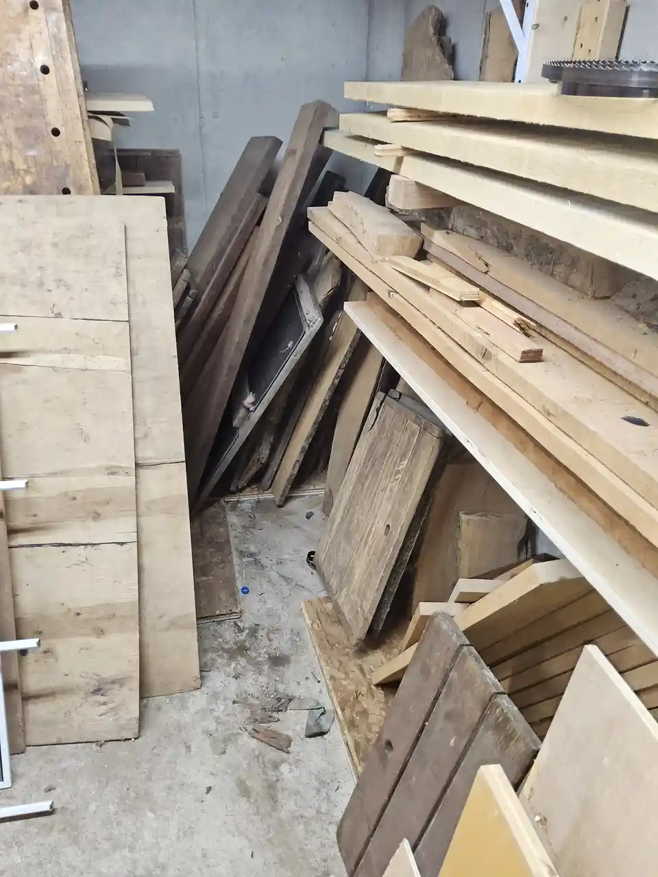 A cluttered workshop or storage room is filled with stacks of lumber, plywood, and wooden planks of various sizes and…