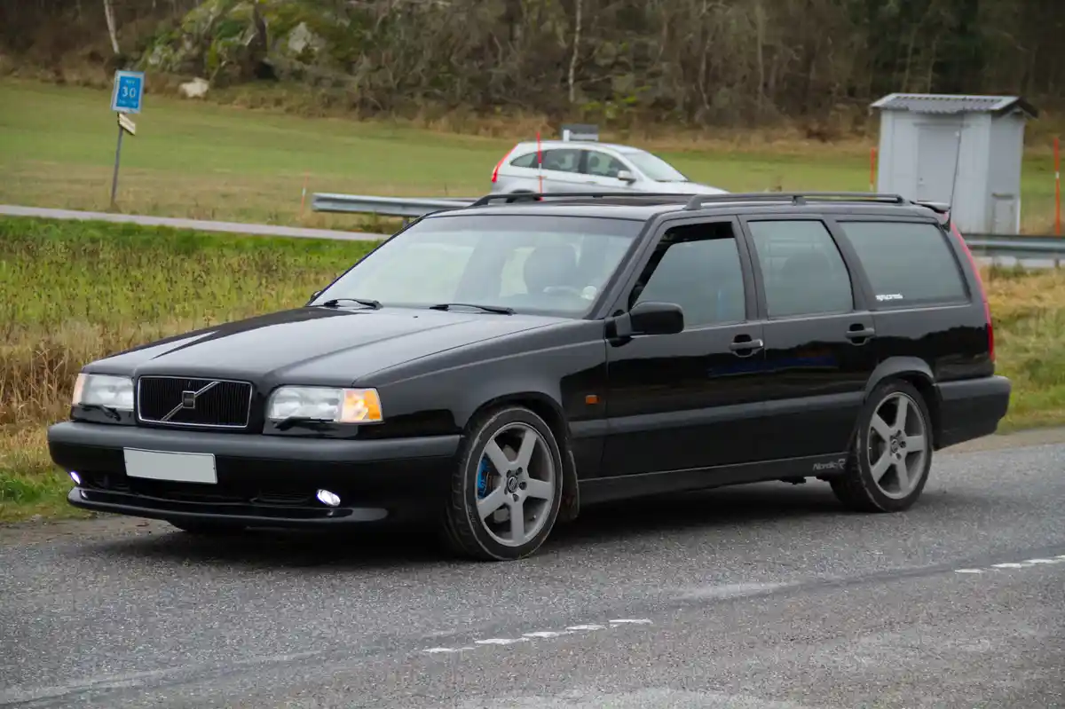 A black Volvo V70 R station wagon is parked on an asphalt road with a grassy field and trees in the background. A blue speed…