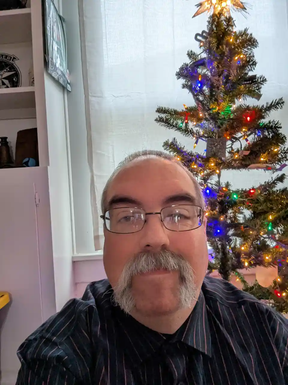 A man with glasses and a gray mustache looks directly at the camera with a neutral expression, standing in front of a…