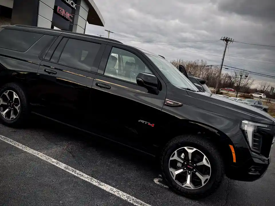 A black GMC Yukon AT4 SUV is parked in a dealership lot under an overcast sky, with the dealership's sign visible in the…