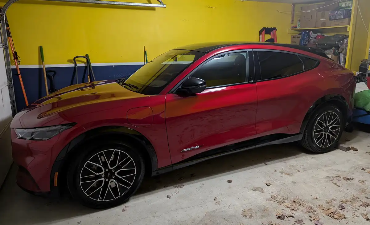 A red Ford Mustang Mach-E electric SUV is parked inside a cluttered garage with yellow and blue walls, surrounded by tools…