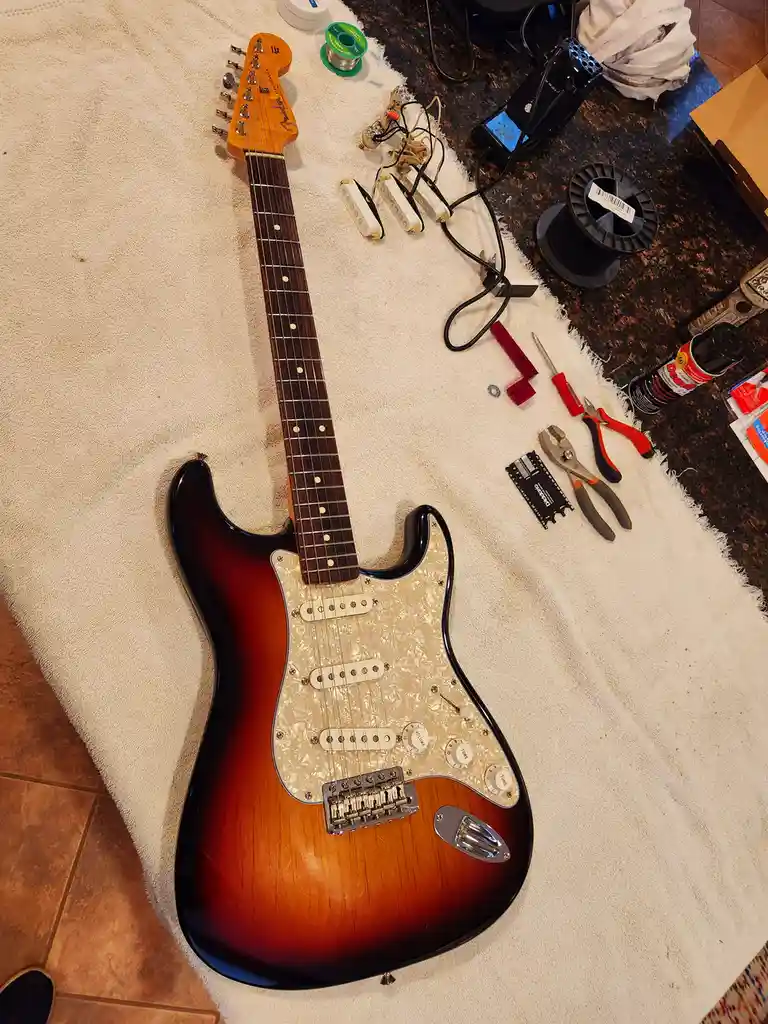 An electric guitar with a sunburst finish lies on a white towel, surrounded by tools and parts like pickups, solder, pliers…