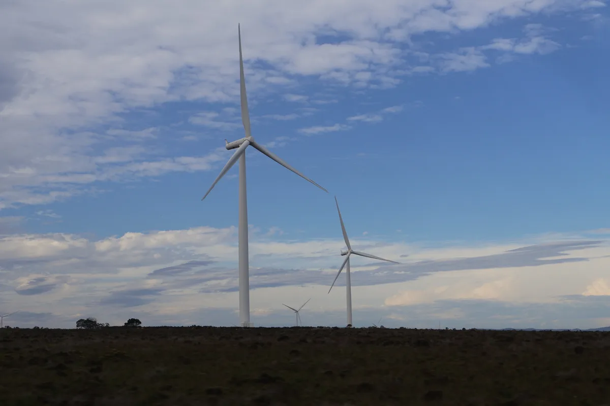 A landscape photograph shows several large white wind turbines standing in a field under a partly cloudy blue sky. The…