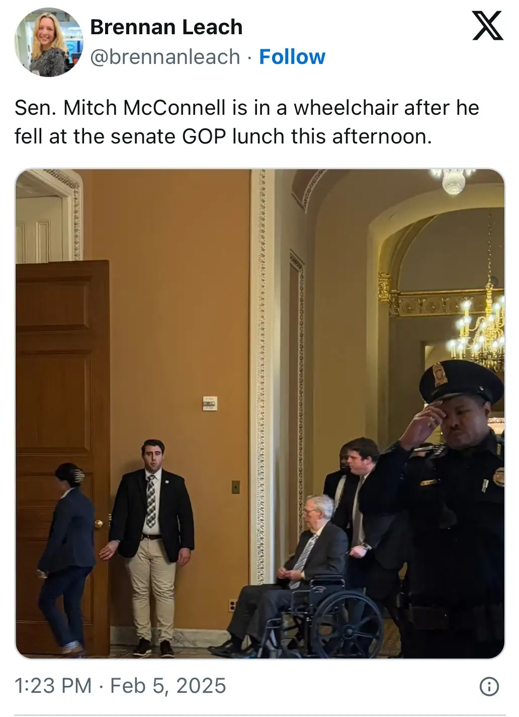 A photo shows an older man in a suit sitting in a wheelchair in an ornate hallway, flanked by several men in suits and a…