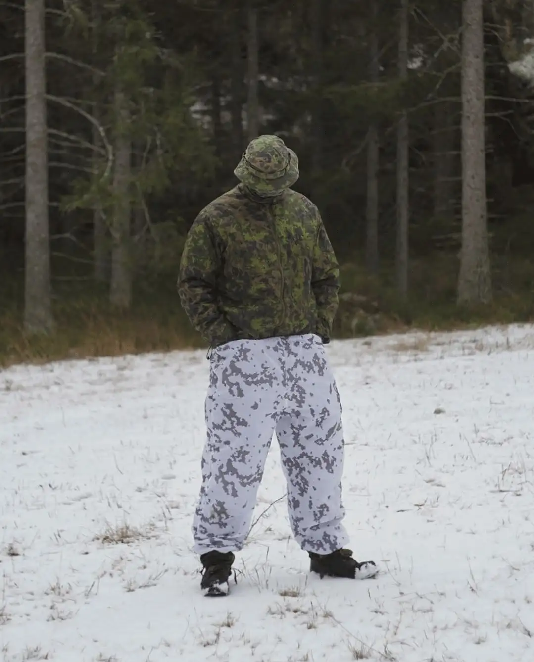 A person stands in a snowy field wearing a green camouflage jacket and hat, but their pants are white with grey digital…