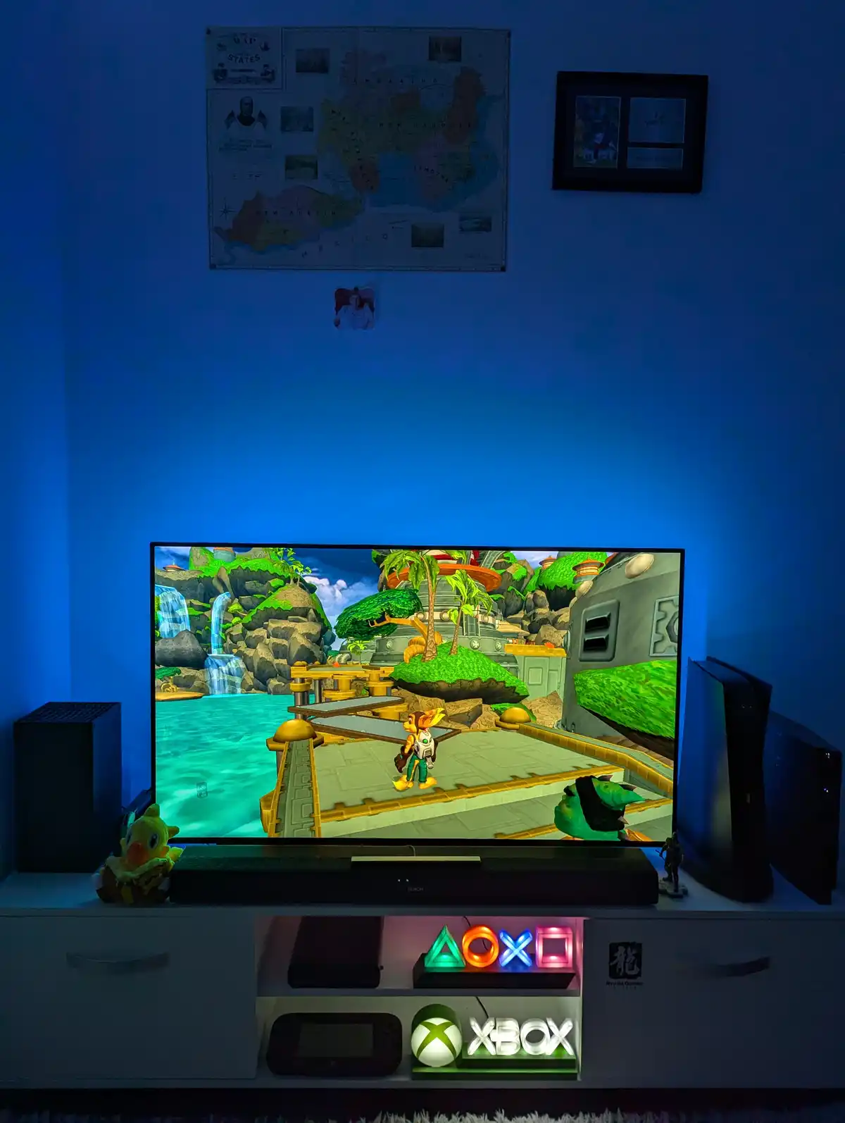 A dimly lit room with blue ambient lighting features a TV displaying a colorful video game scene from 'Ratchet & Clank'…