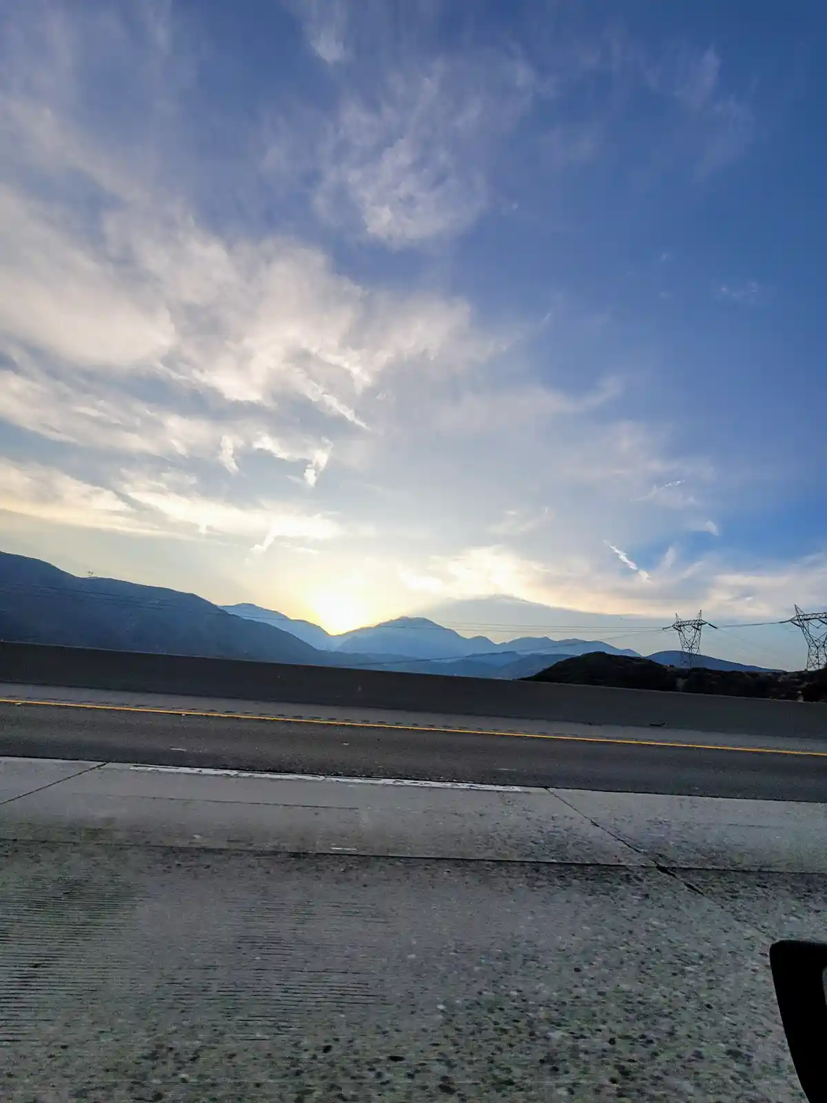 A scenic photo taken from a car window shows a highway with a concrete barrier, distant mountains, power lines, and a bright…