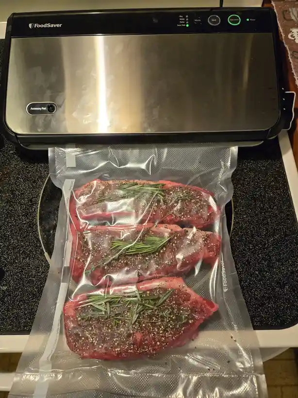 Three raw, seasoned steaks with rosemary and pepper are in a vacuum-seal bag being processed by a FoodSaver machine. The…