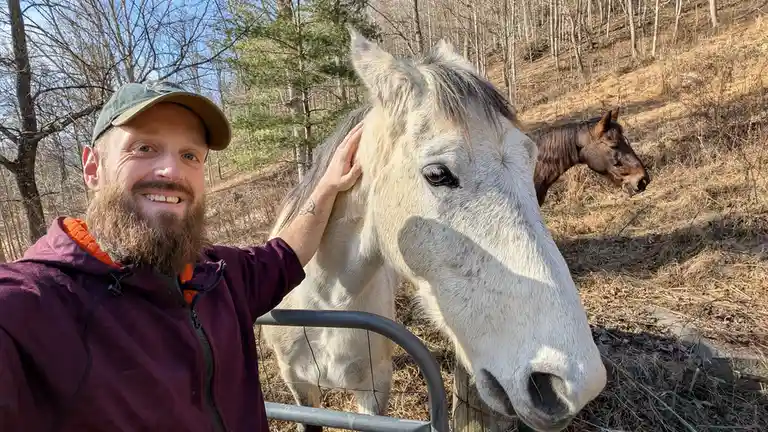 A smiling bearded man in a cap and purple jacket takes a selfie while petting a white horse's head near a fence. A second…