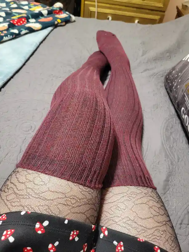 A close-up photo shows a person's legs resting on a gray bedspread, wearing ribbed burgundy tights over black patterned…