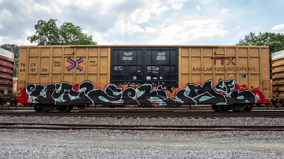 A tan freight railcar with 'railcar pooling experts' and 'ttx' branding sits on tracks, covered in large black and teal…