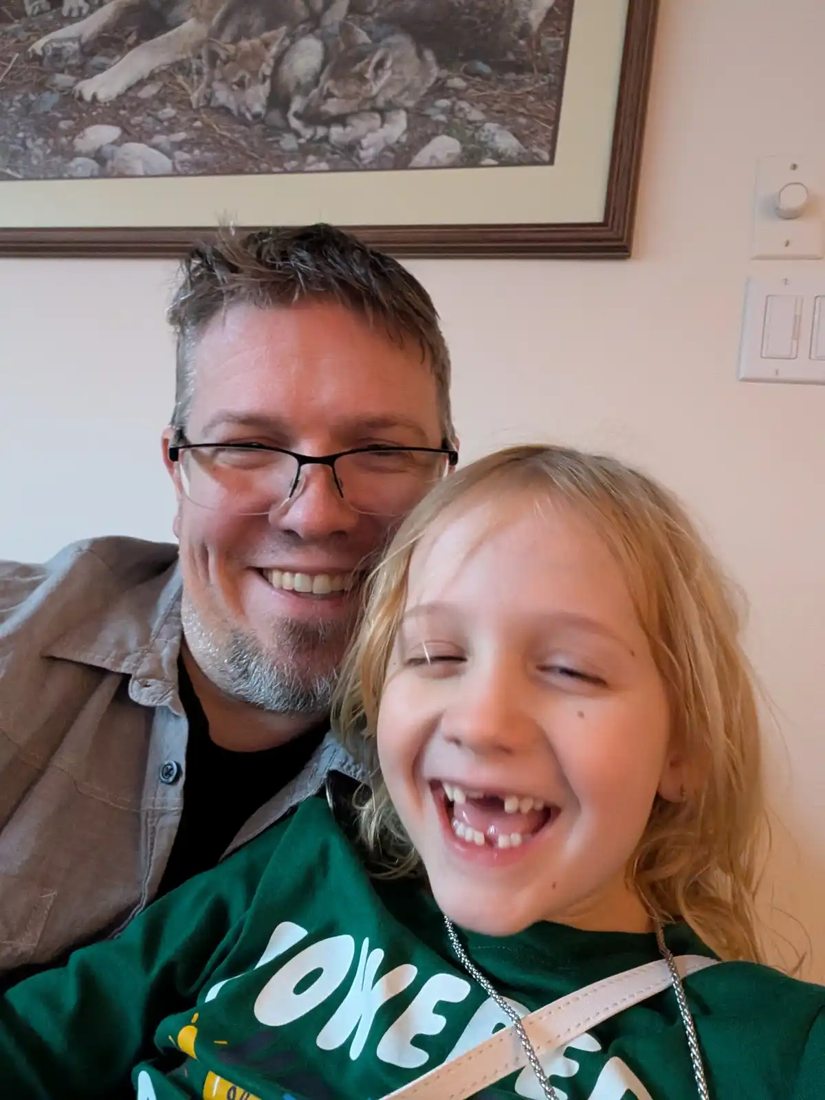 A man and a young girl are smiling and laughing closely together in a selfie. Behind them, a framed picture on the wall…