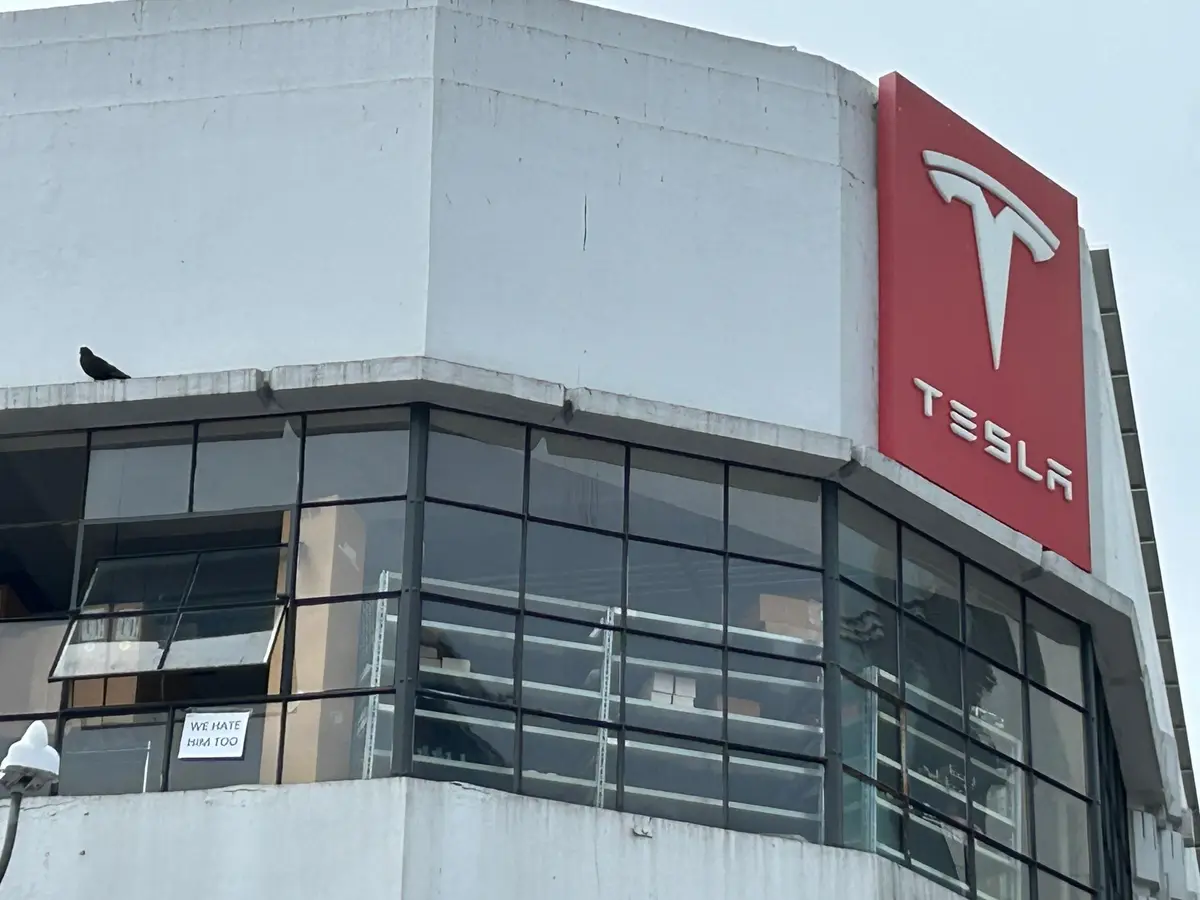 A photo of a Tesla building with its red logo and white 'TESLA' text. A small white sign taped to a window reads 'WE HATE…