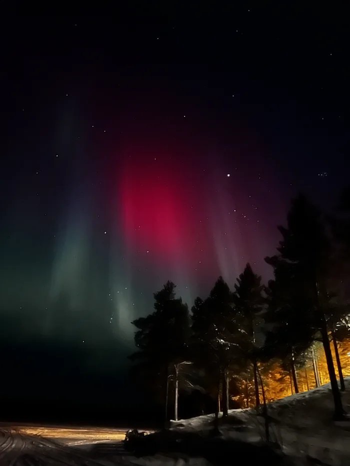 A nighttime photograph shows a vibrant red and green aurora borealis dancing across a starry sky above a snowy landscape…