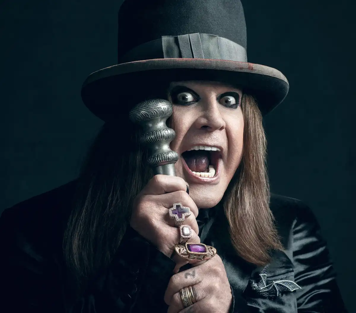 A man with long brown hair, heavy black eye makeup, and a black top hat holds a silver cane to his face while screaming with…