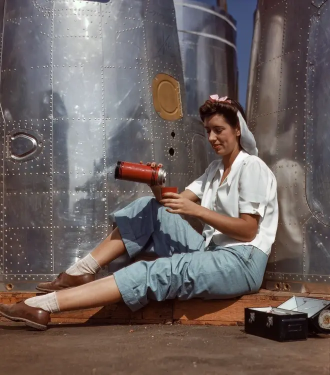 a woman in a white shirt and blue pants sits on a wooden plank against large silver airplane fuselage sections, pouring from…
