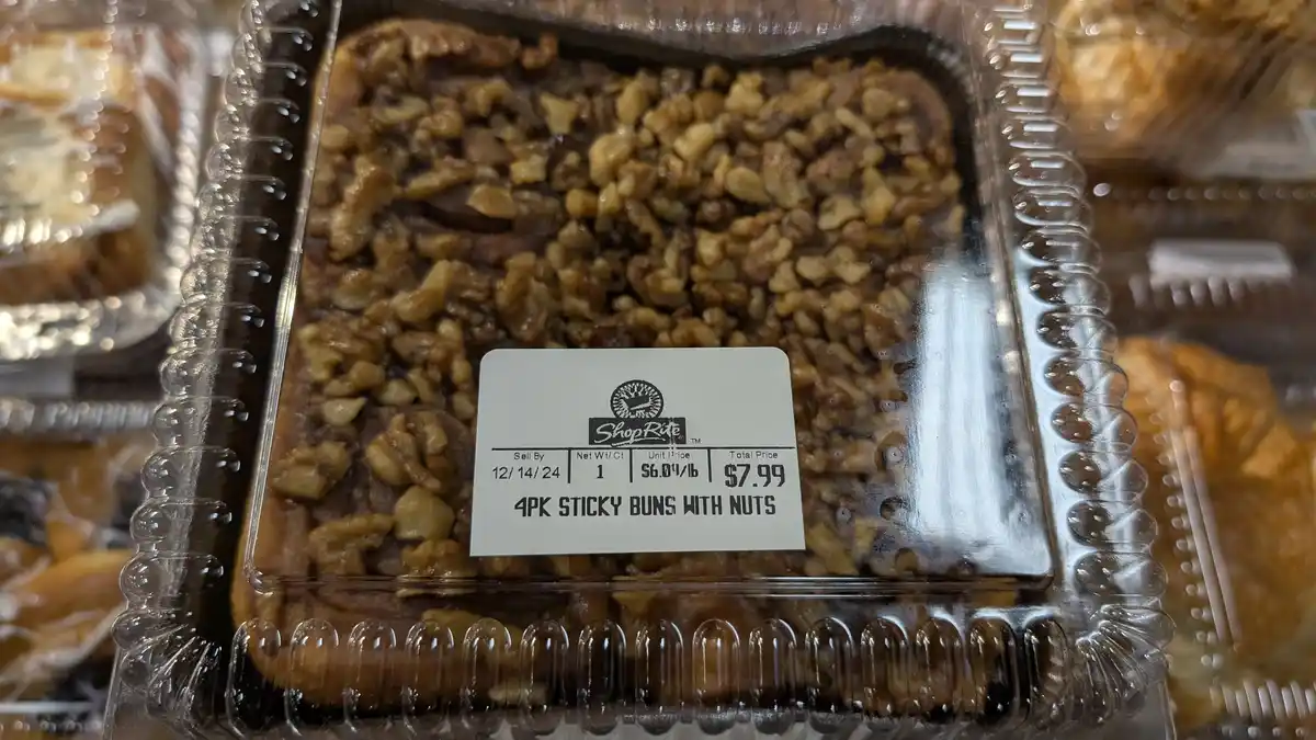 A close-up photo of a clear plastic bakery container holding four sticky buns topped with chopped walnuts. The white…