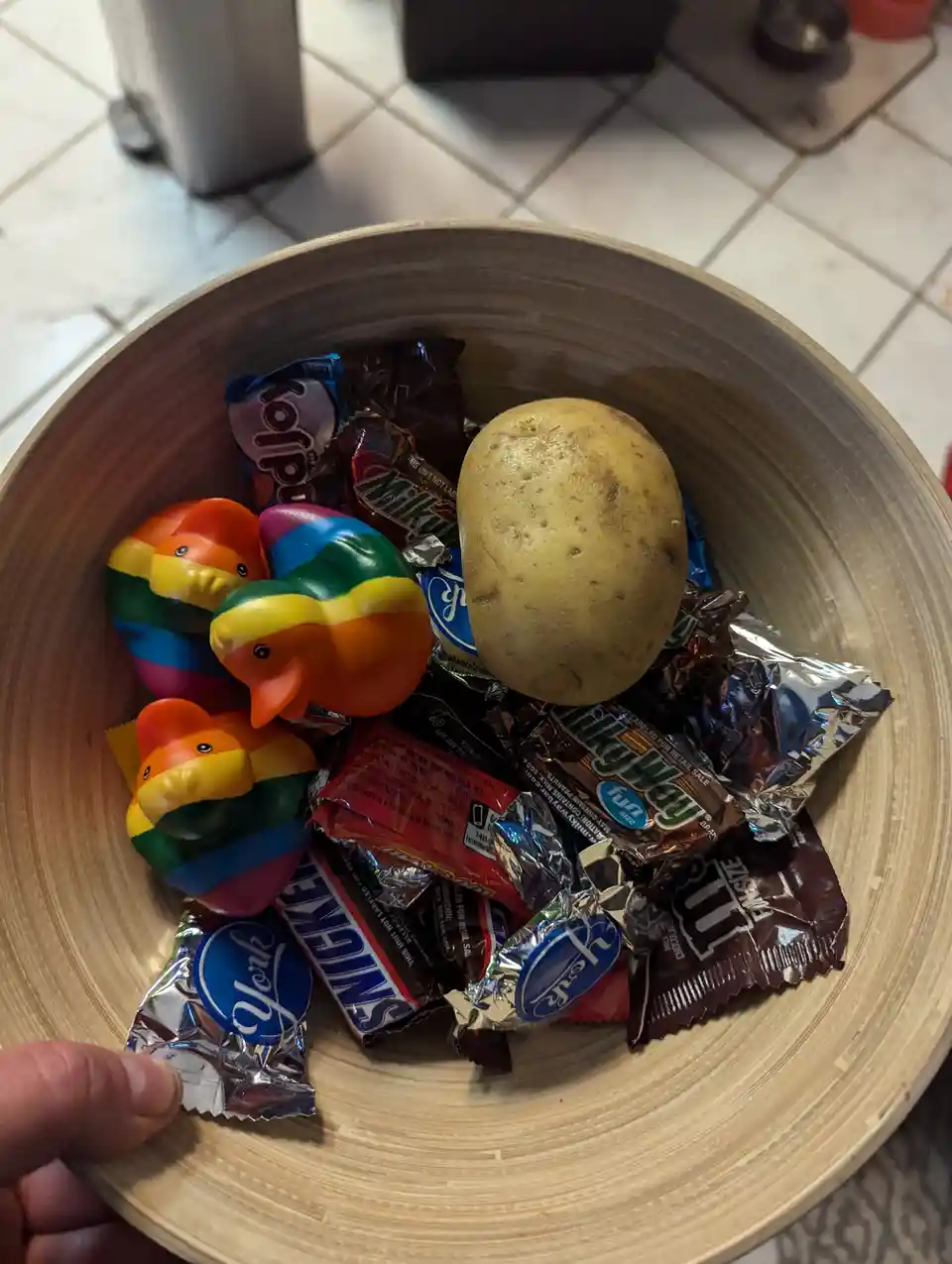 A wooden bowl filled with various wrapped chocolate candies and three small rainbow-colored rubber ducks, with a single…