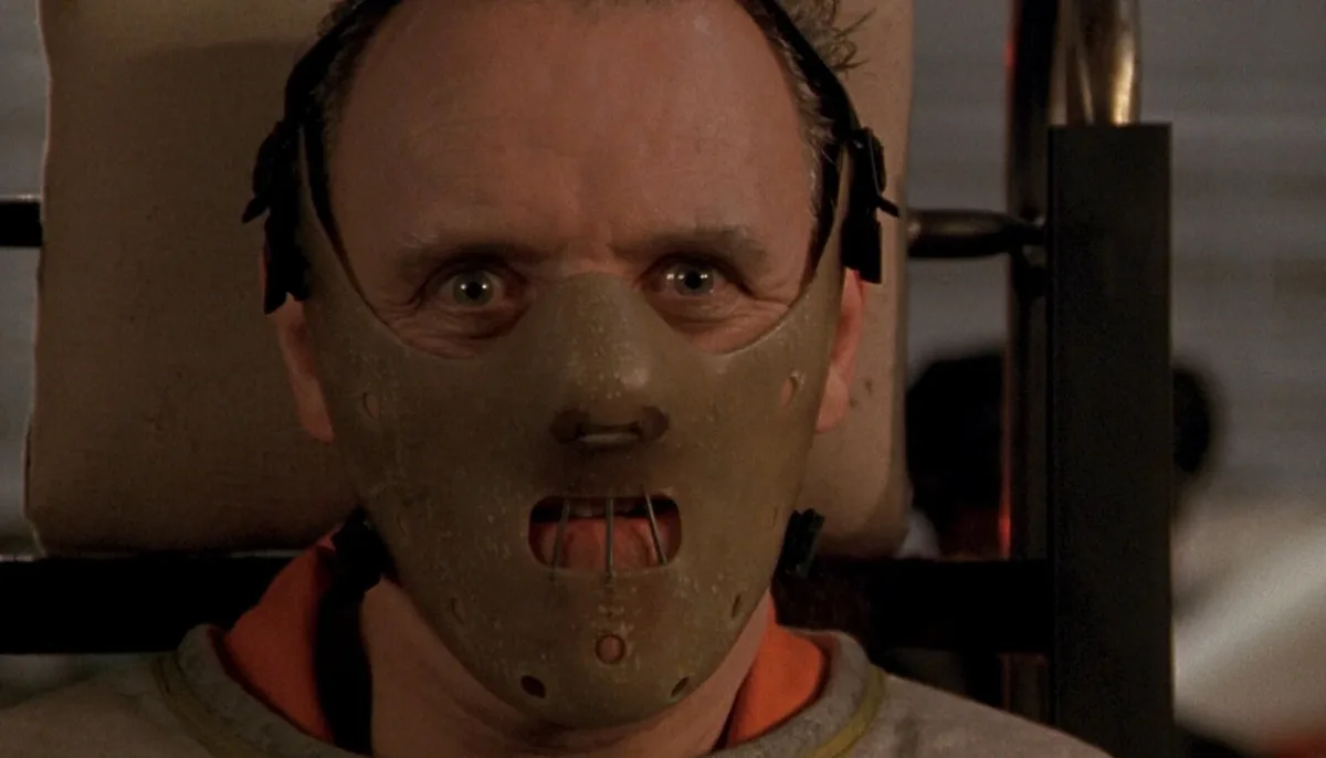 A man with wide, terrified eyes is strapped into a chair, wearing a grey, textured mask that covers his nose and mouth, with…