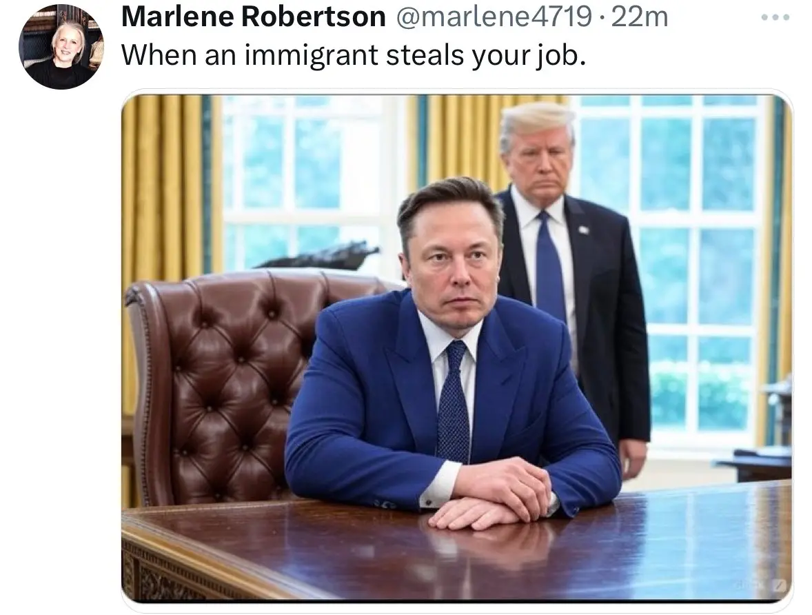 A man in a blue suit sits at a large wooden desk with his hands clasped, looking serious. Behind him, another man in a dark…