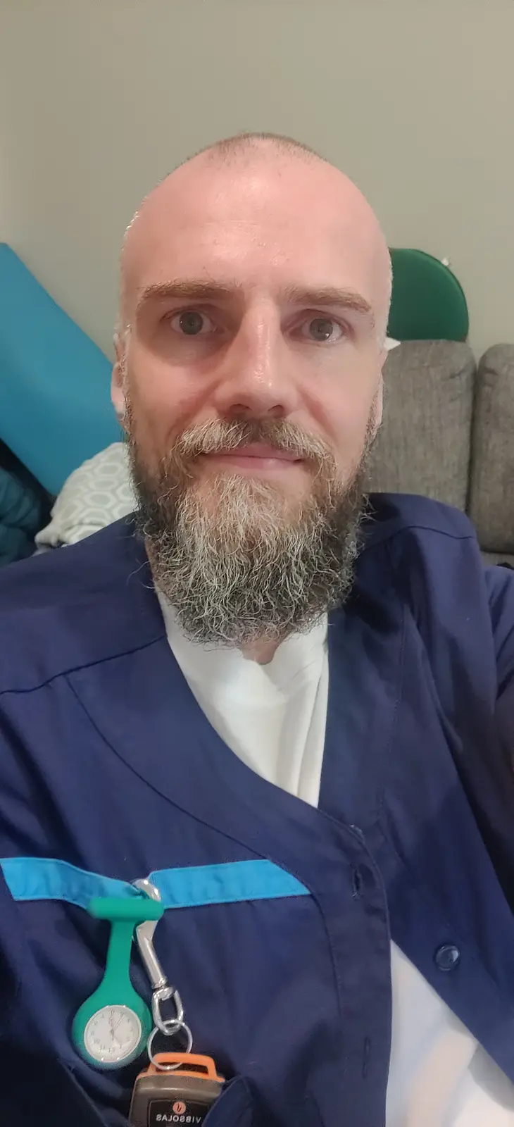 A man with a graying beard and shaved head, wearing blue medical scrubs over a white shirt, sits in a chair. A green…