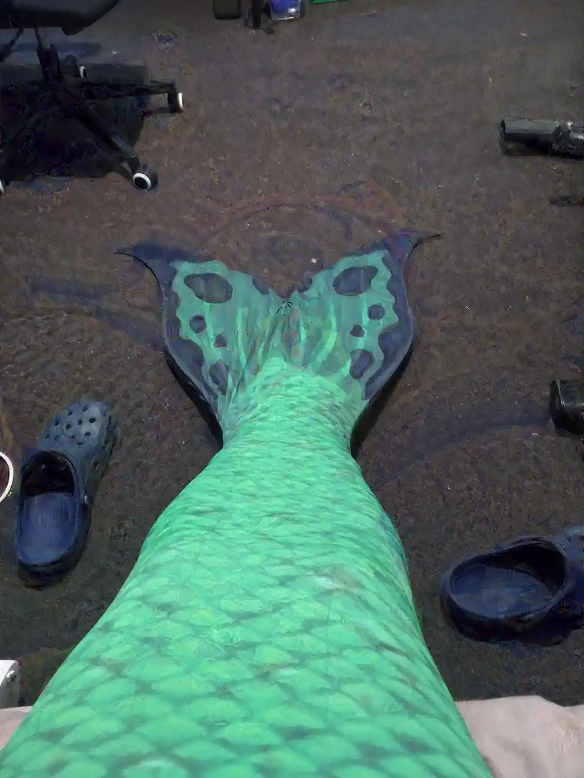 A top-down photo shows a person wearing a green, scale-patterned mermaid tail costume lying on a dark floor, with black…