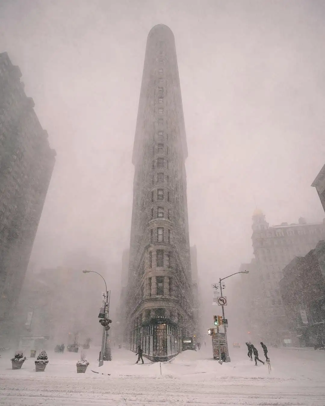 A snowy, atmospheric photo of the Flatiron Building in New York City, with heavy snowfall obscuring the background and a few…