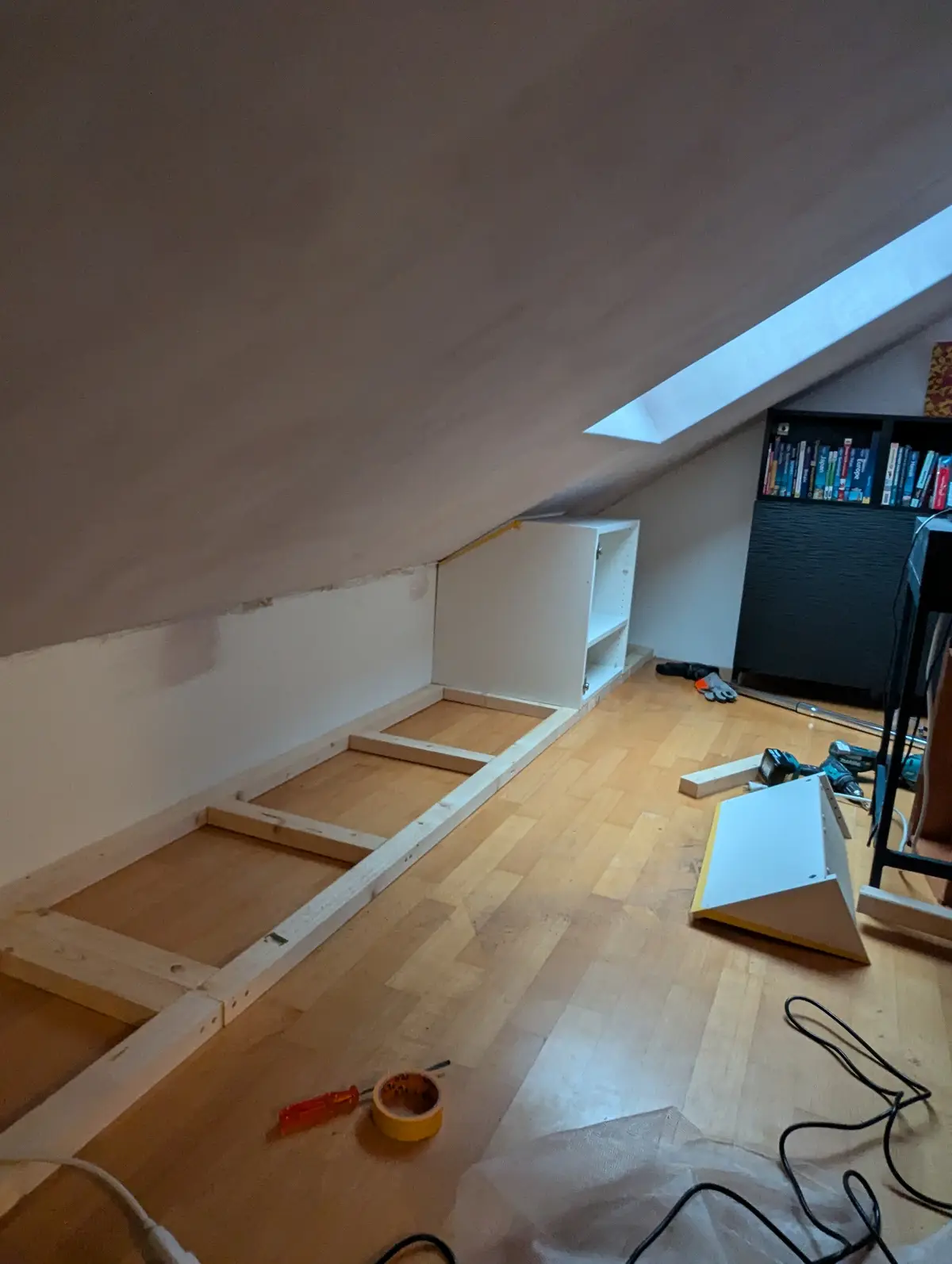 A room with a slanted ceiling and a skylight shows a partially assembled white IKEA bed frame lying on the wooden floor…