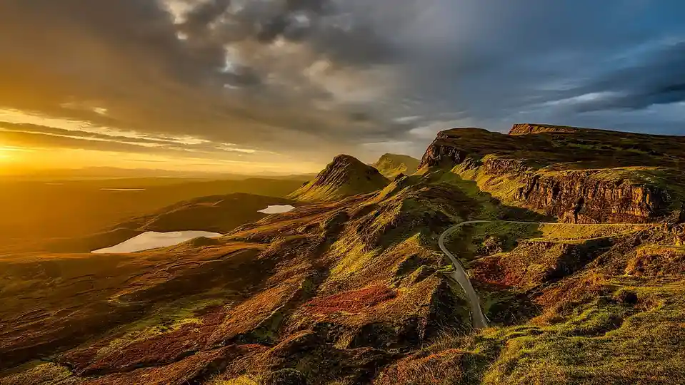 A scenic landscape photo of a winding road on a mountainous terrain at sunset, with golden light illuminating the hills and…