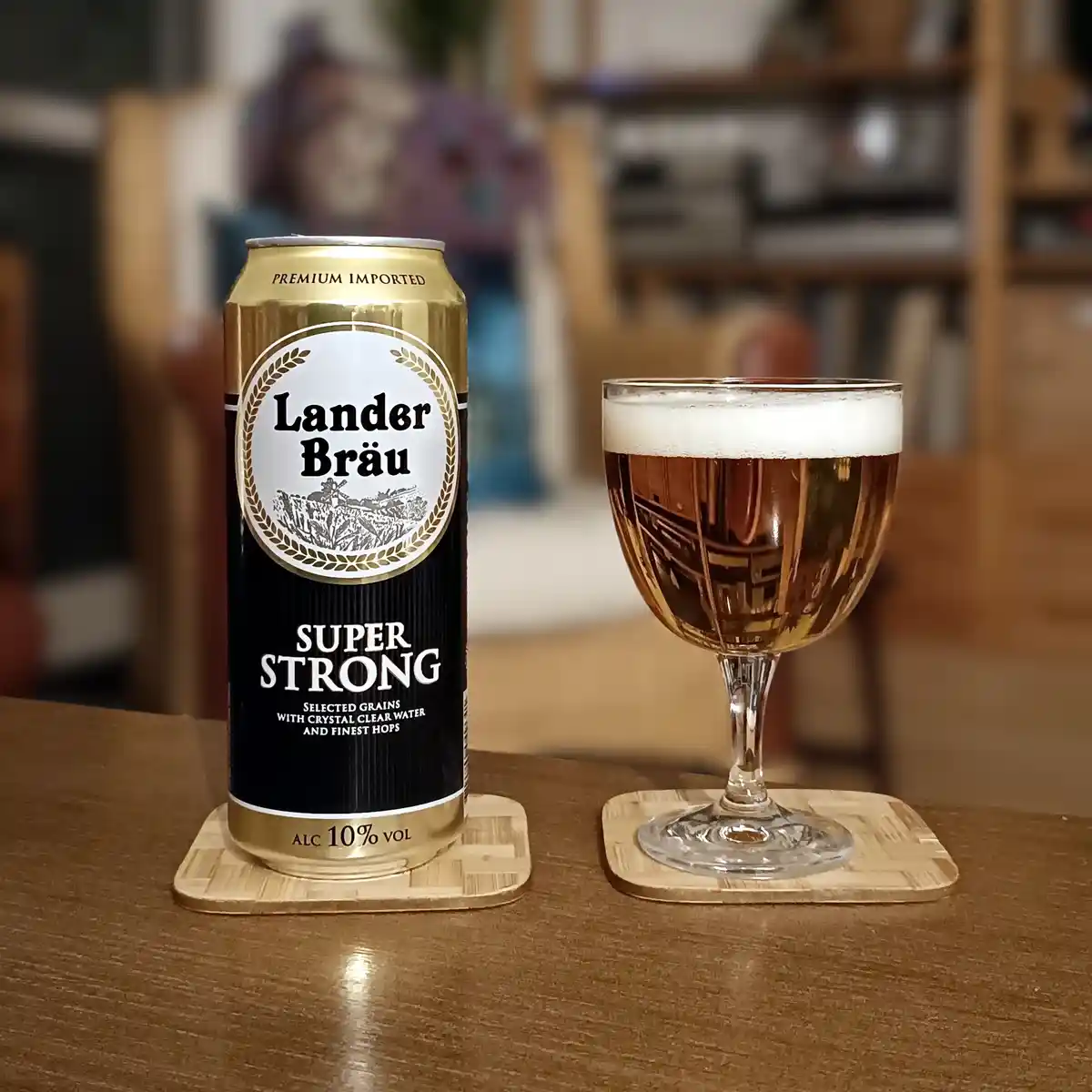 A gold and black can of Lander Bräu Super Strong beer, labeled with 10% alcohol, sits next to a stemmed glass filled with…