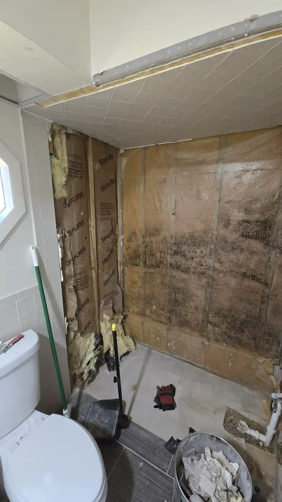 A bathroom under renovation shows exposed insulation and damaged walls, with a toilet still intact in the corner. Tools and…