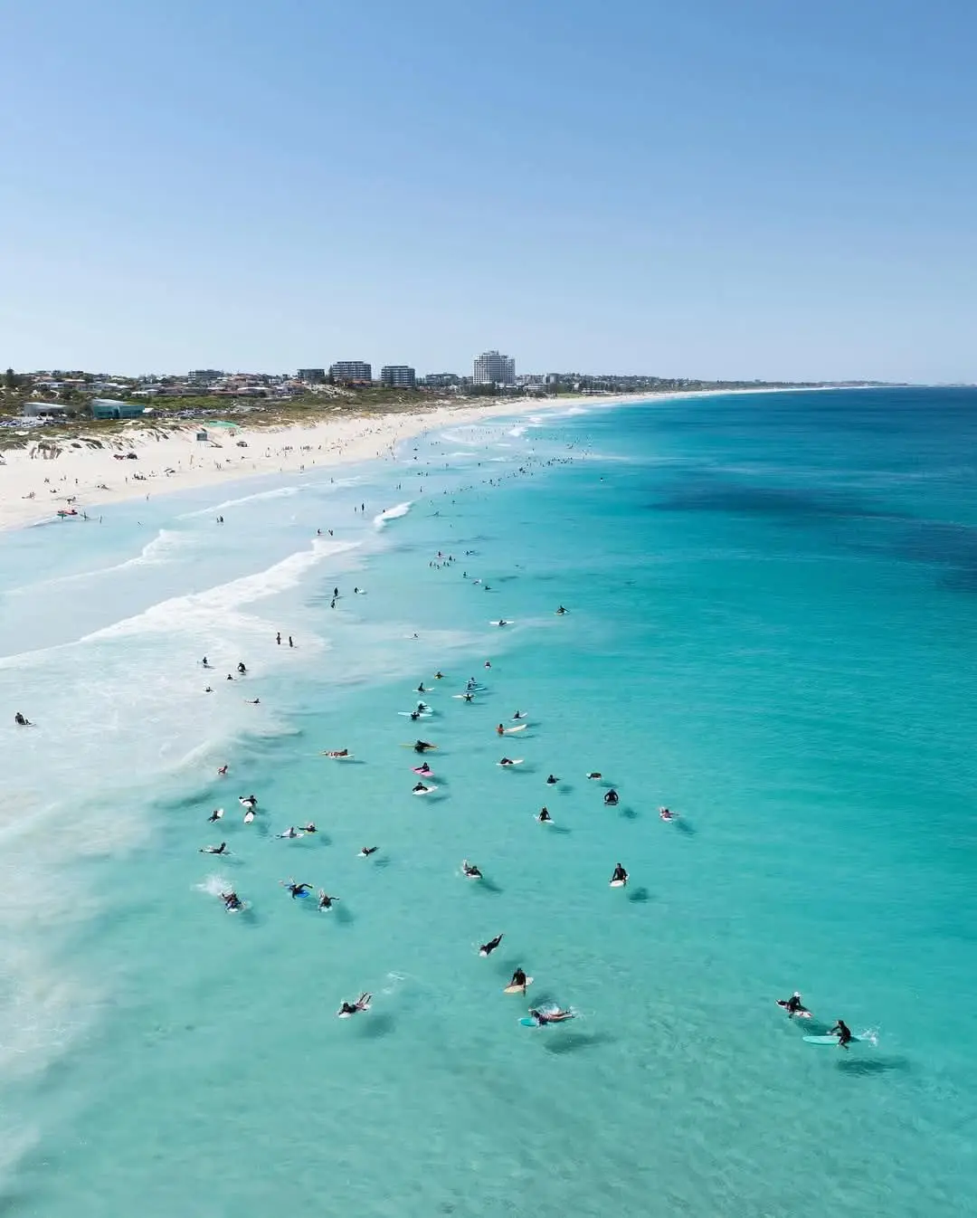 An aerial photo shows a crowded beach with many surfers in the turquoise water and people on the white sand, under a clear…