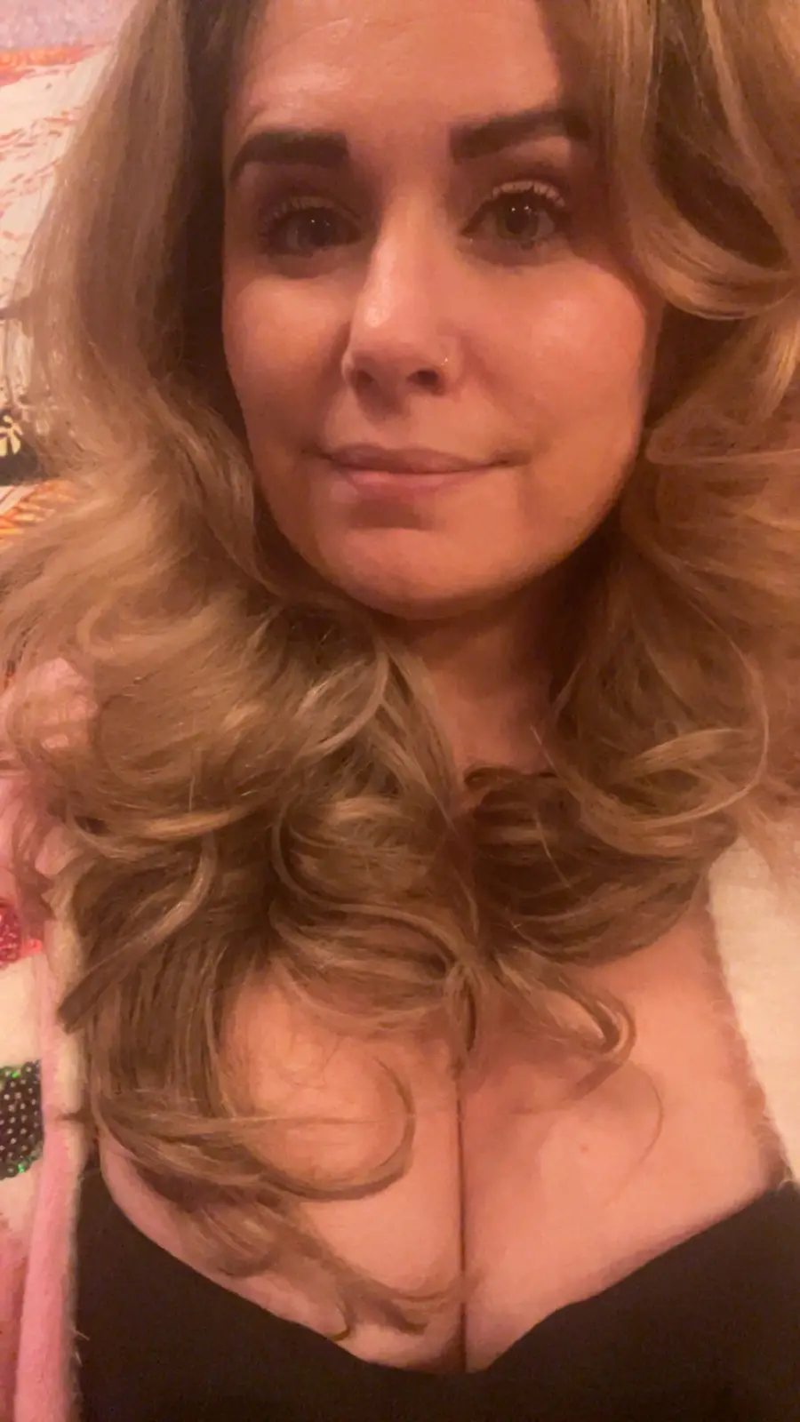 A close-up selfie of a woman with long, wavy blonde hair, wearing a black top and a pink fuzzy cardigan with sequins. She…