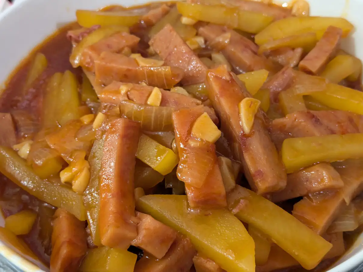 A close-up photo of a bowl of stew-like food containing chunks of pink processed meat, yellowish potato-like pieces, and…