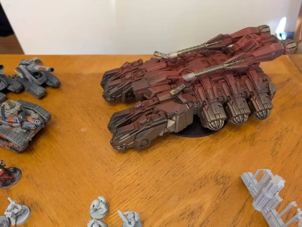 A large, heavily detailed red and brown painted sci-fi tank model sits on a wooden table, surrounded by smaller unpainted…