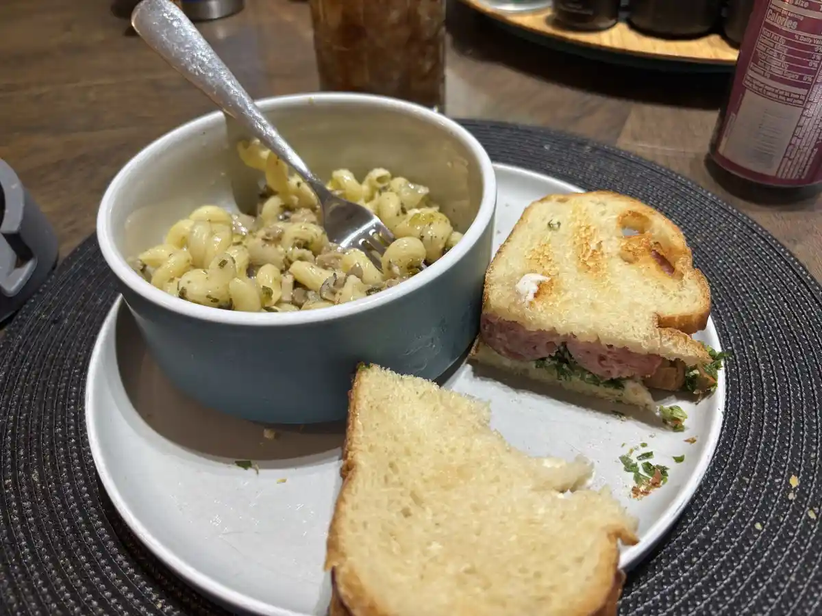 A plate holds a partially eaten sandwich with visible meat and greens, alongside a bowl of macaroni and cheese with a fork…
