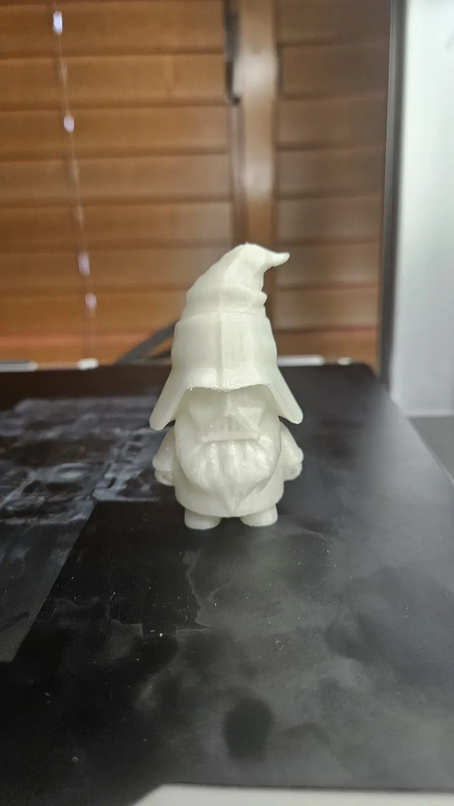 A white 3d-printed figurine of a wizard with a disproportionately large, floppy hat sits on a black surface. The hat covers…