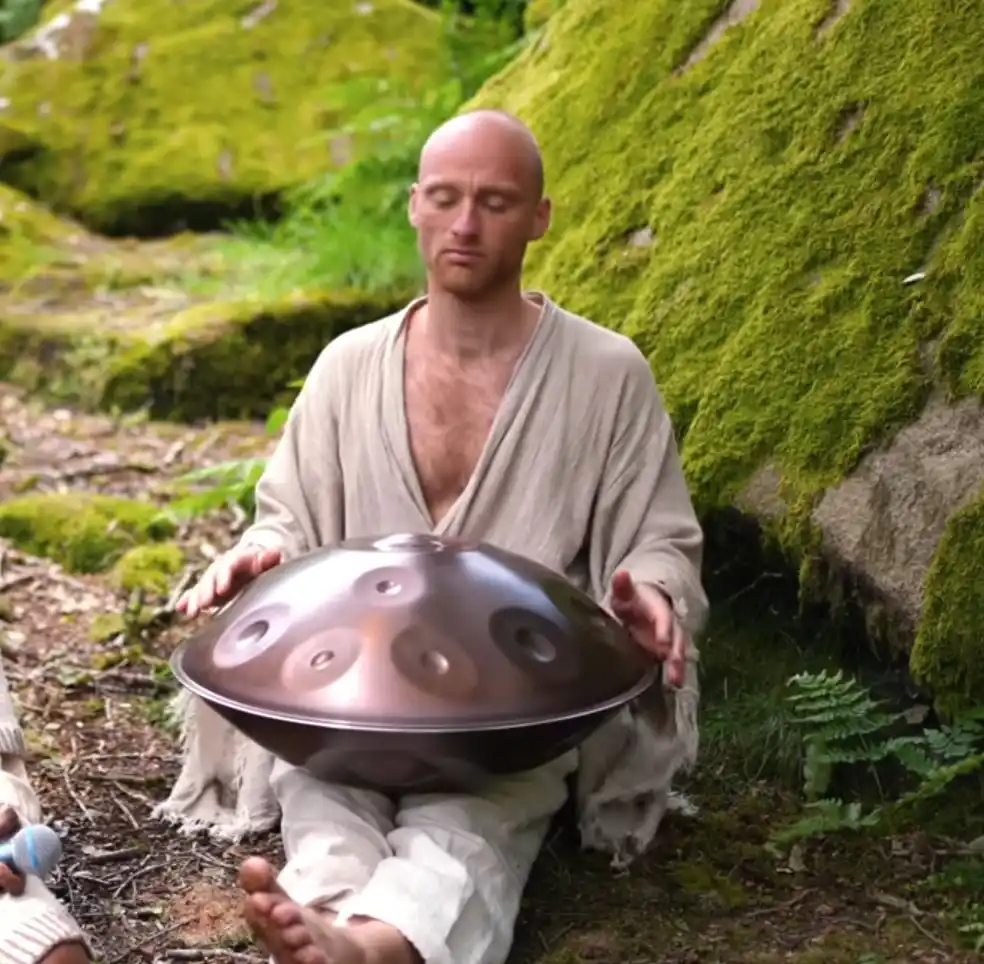 A bald man in loose beige robes sits cross-legged on mossy forest ground, holding a handpan (a metallic, bowl-shaped…