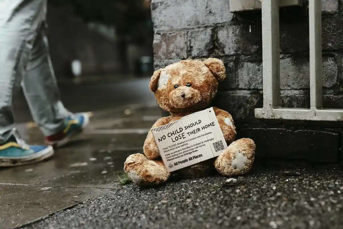 A worn, patched brown teddy bear sits alone on a wet sidewalk against a brick wall, holding a cardboard sign with text about…