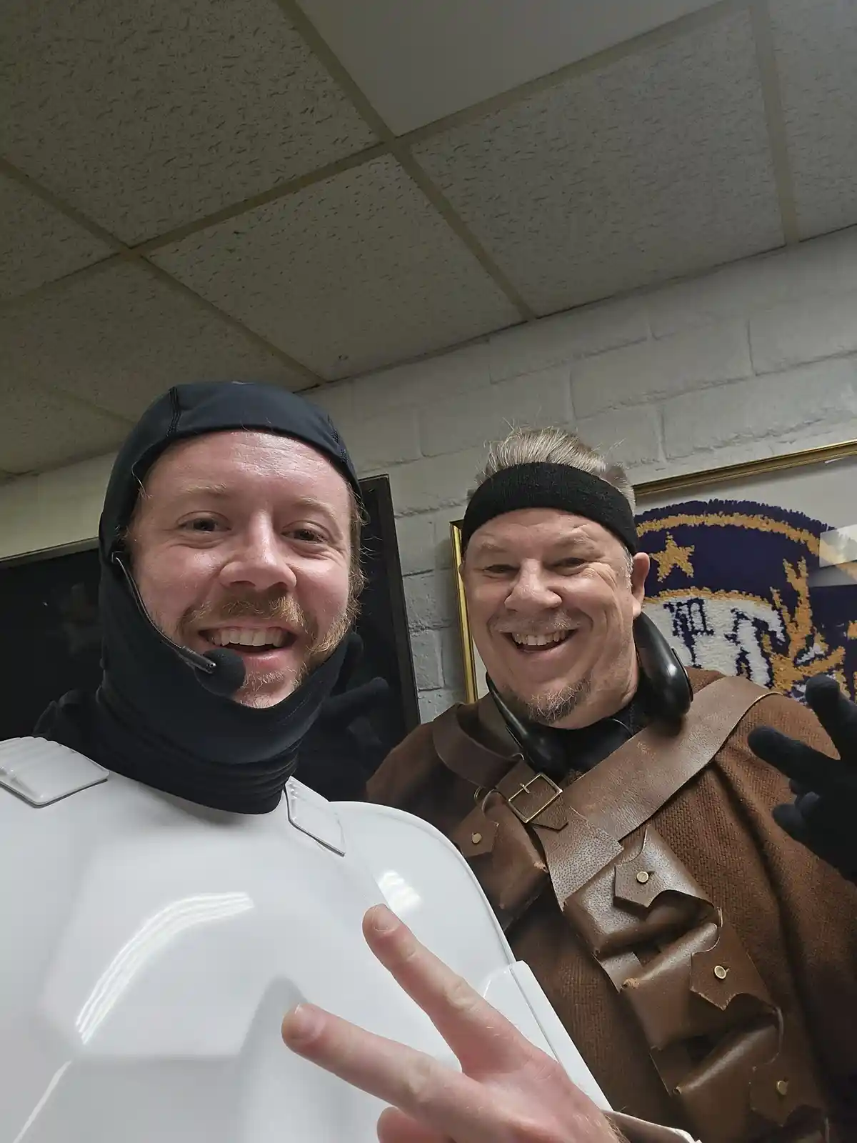 Two men in costumes pose for a selfie in an office-like setting. The man on the left wears a white stormtrooper helmet and a…