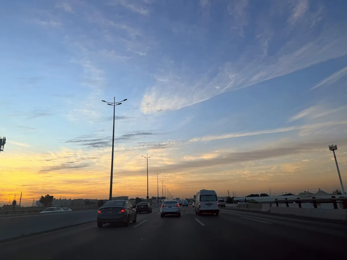 A photo taken from inside a car on a highway during sunset, showing multiple cars ahead under a sky with orange and blue…