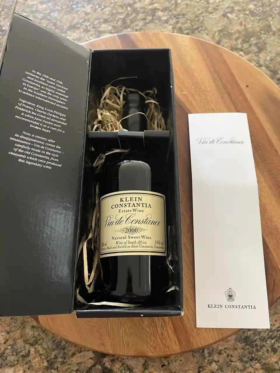A bottle of Klein Constantia Vin de Constance 2000 wine sits in its black presentation box, which has text claiming…