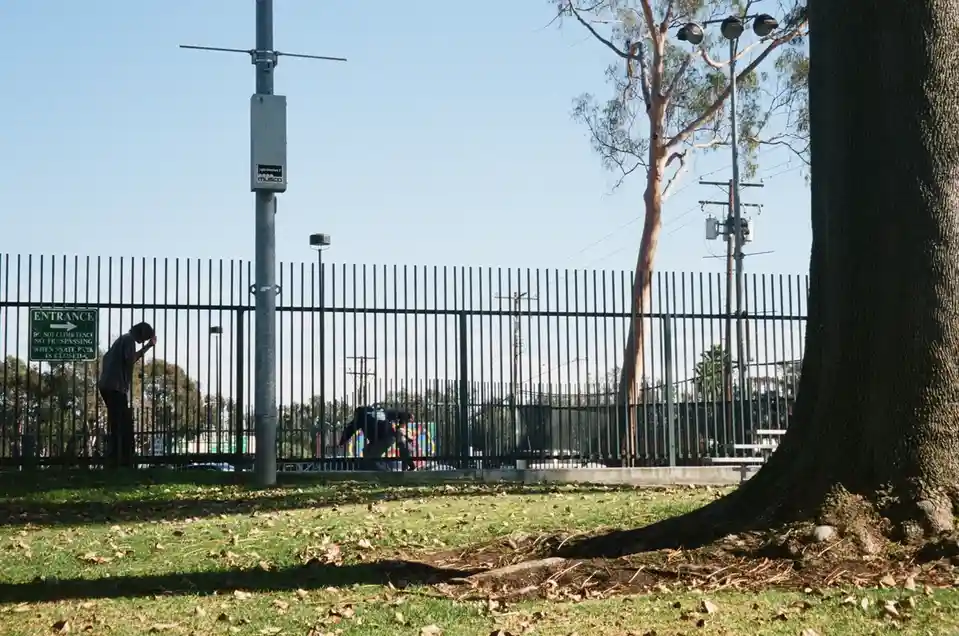 A person leans against a tall metal fence at a skate park entrance, with a green sign reading 'ENTRANCE DO NOT CLIMB FENCE…