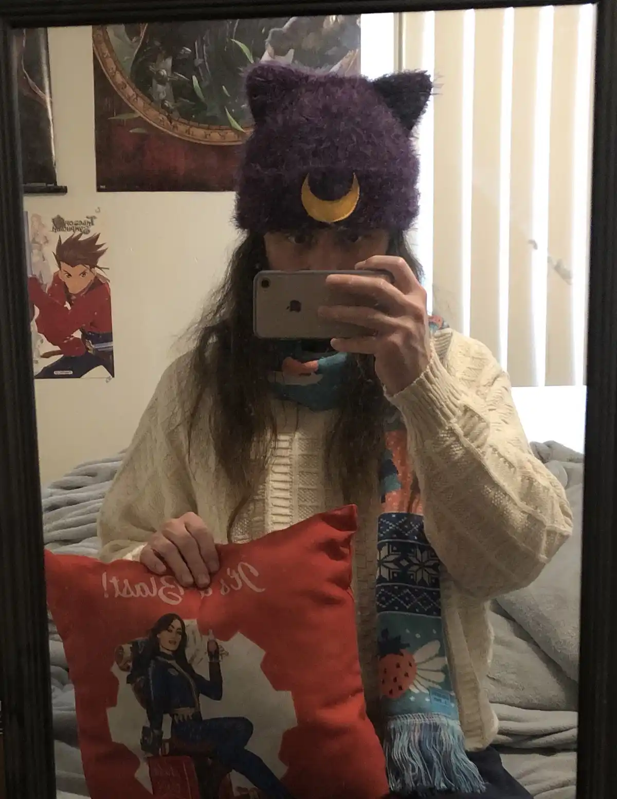 A person with long hair takes a mirror selfie while wearing a purple cat-ear hat with a yellow crescent moon emblem, a…
