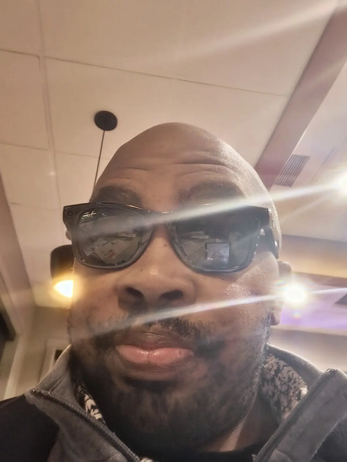 A close-up selfie of a bald man with a beard wearing dark sunglasses, taken indoors under bright, glaring lights that create…