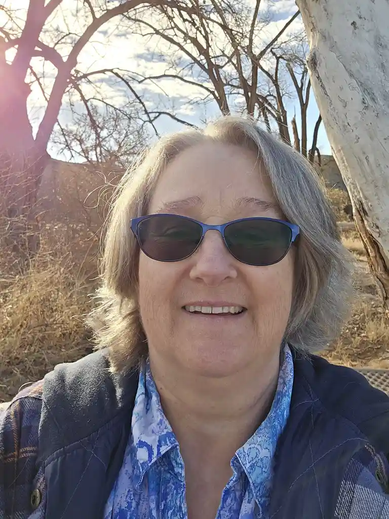 A woman with gray hair and blue sunglasses smiles at the camera while sitting outdoors under bare trees with sunlight…