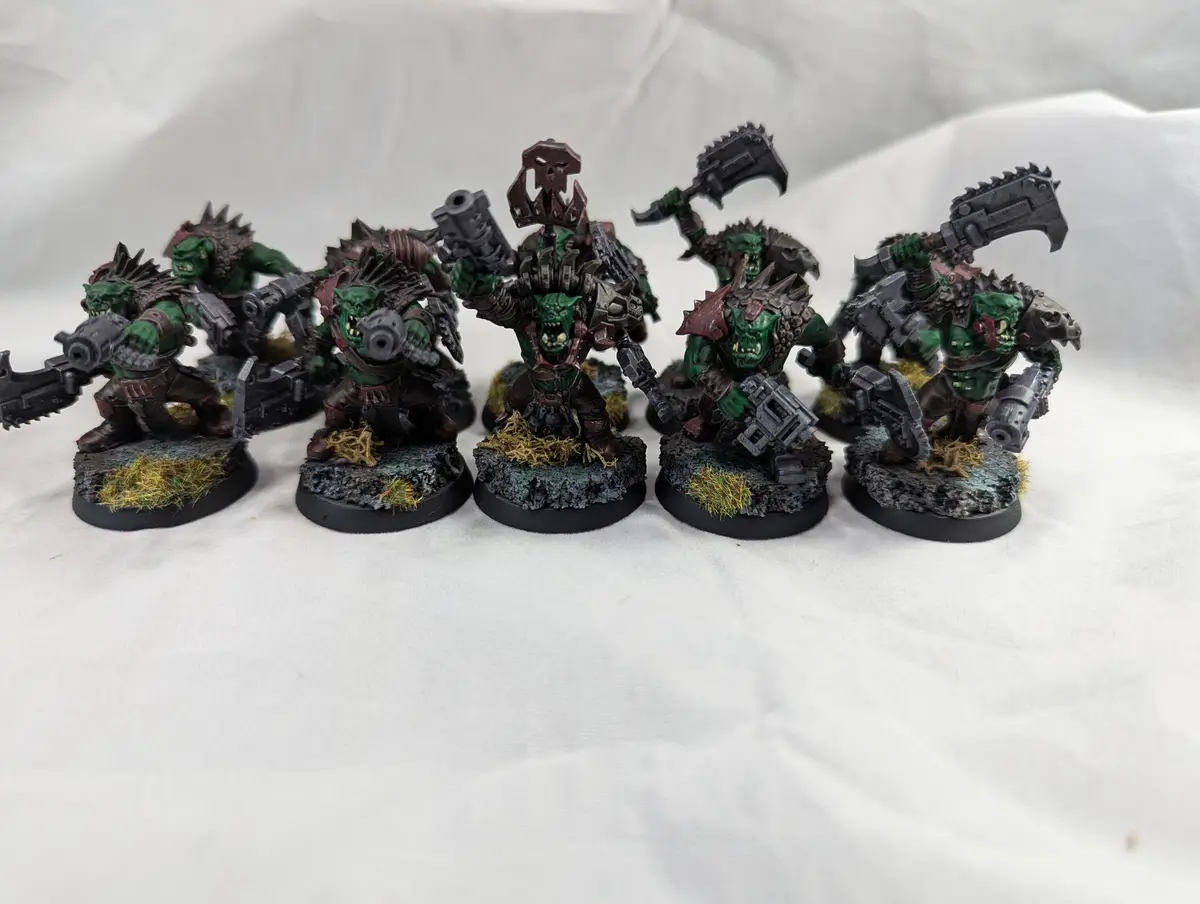 A row of six green-skinned, heavily armed fantasy ork miniatures, each standing on a textured base with grass tufts…