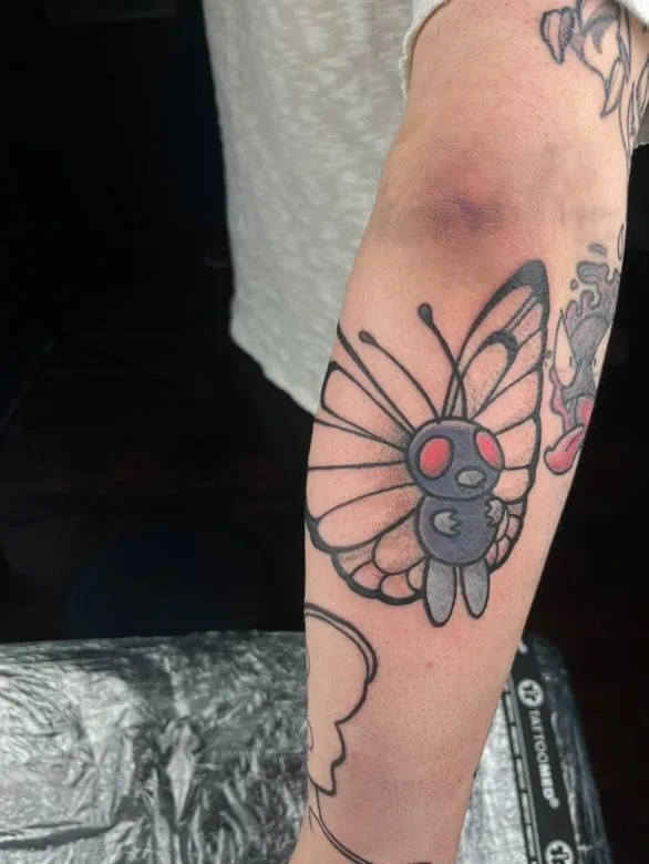 A close-up photo shows a person's forearm with a freshly inked tattoo of a butterfly. The butterfly's body is designed to…