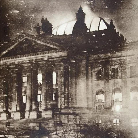 A grainy, black-and-white historical photograph shows a large, ornate building with classical columns and a domed roof…